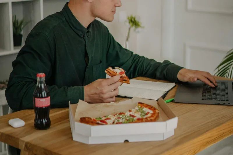 Das Bild zeigt einen jungen Mann, der am Computer Pizza isst und Cola trinkt. Hektisches, unbewusstes Essen sowie mangelndes Kauen können die Verdauung beeinträchtigen und zu vermehrter Luftaufnahme oder Blähungen führen.
